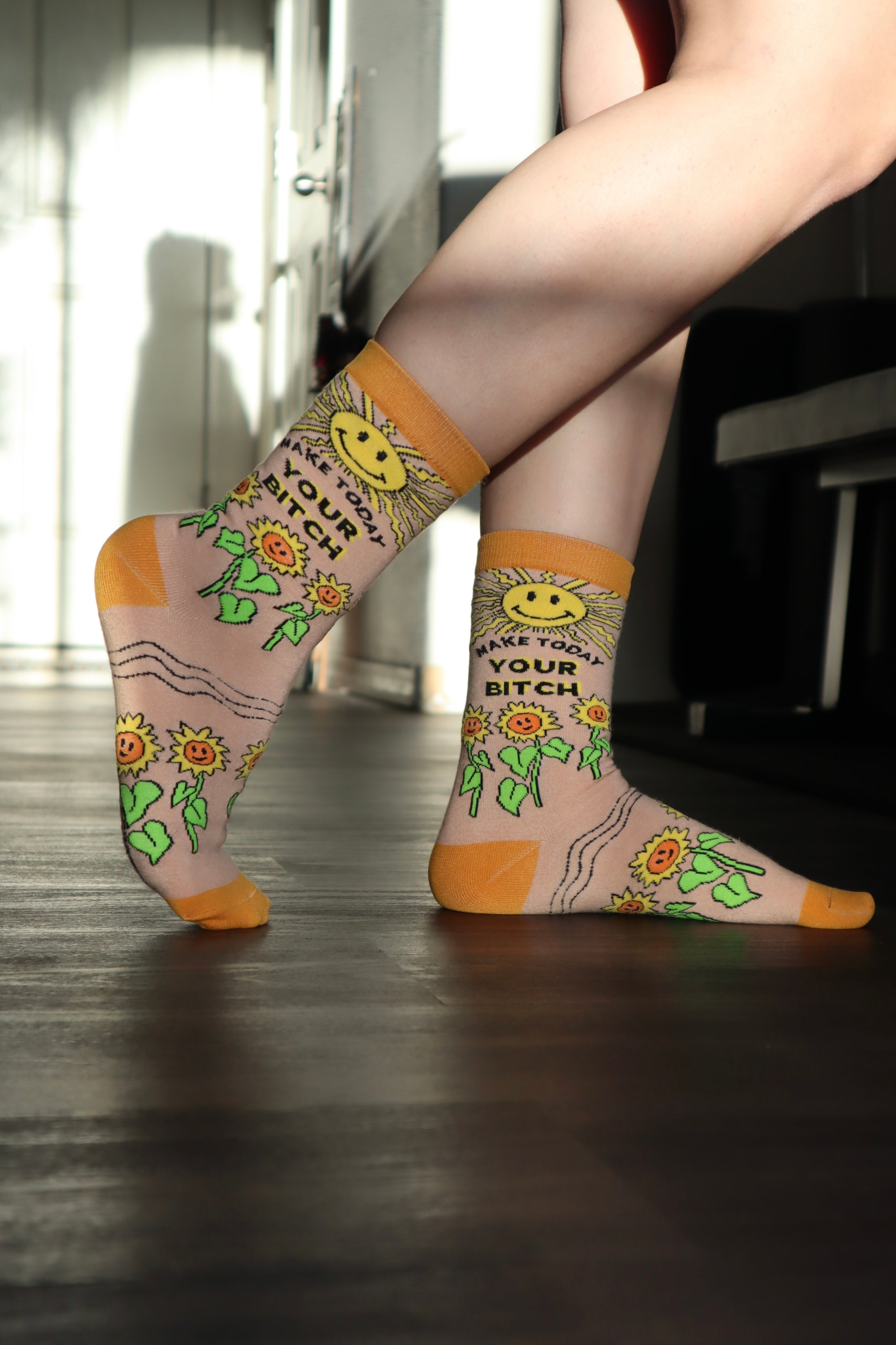 Make Today Your B**tch Socks. Model wearing a pair of socks depicting the sun and sunflowers with a bold sweary slogan. Orange toes, cuff and heel.