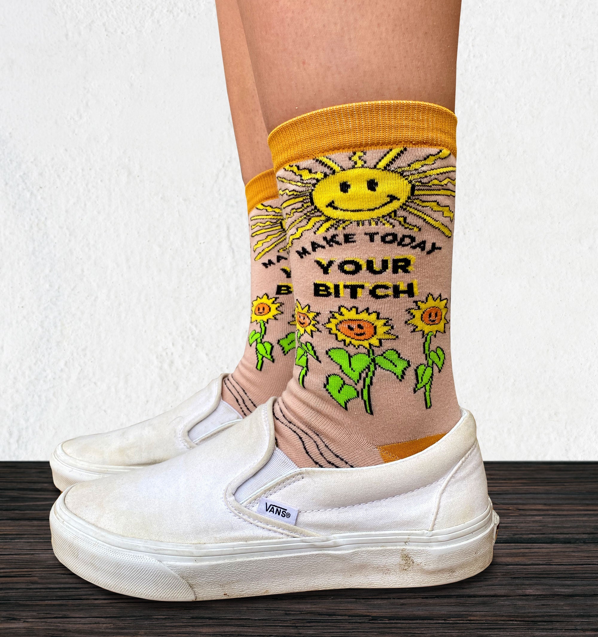 Make Today Your B**tch Socks. Model wearing a pair of socks depicting the sun and sunflowers with a bold sweary slogan. Orange toes, cuff and heel.