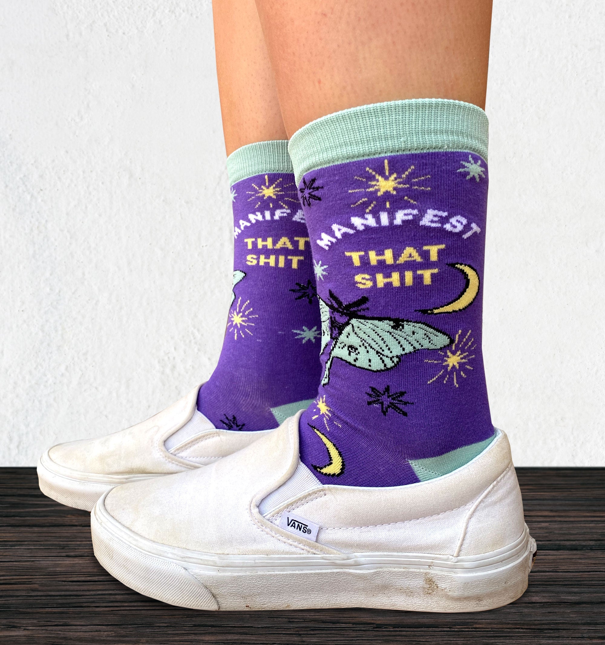 Model wearing a pair of socks with the slogan Manifest That S**t and a butterfly motif. Purple ankles, turquoise toe, heel and cuff.