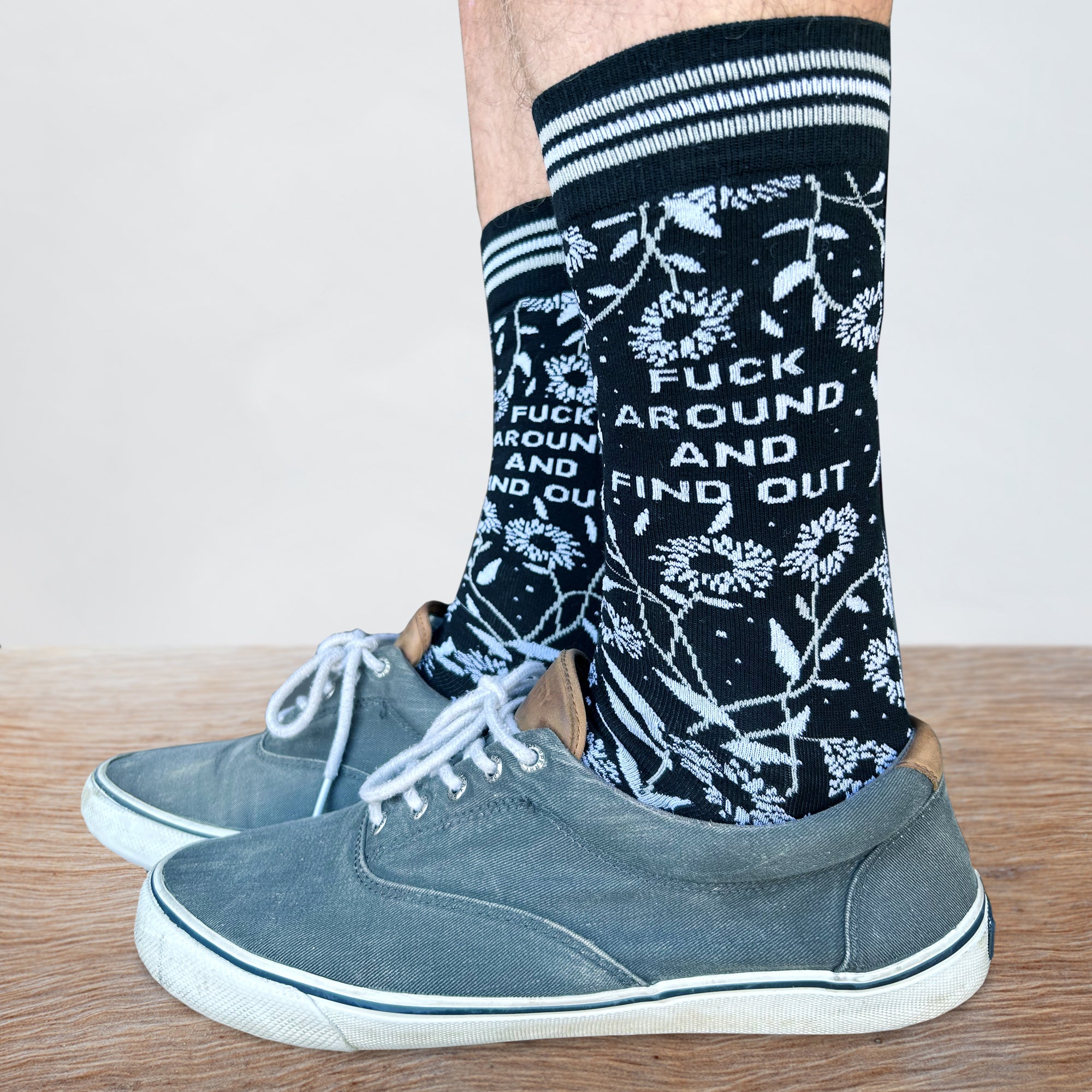 Model wearing a pair of bold floral and slogan socks with black legs and light blue toes, cuff and heel.
