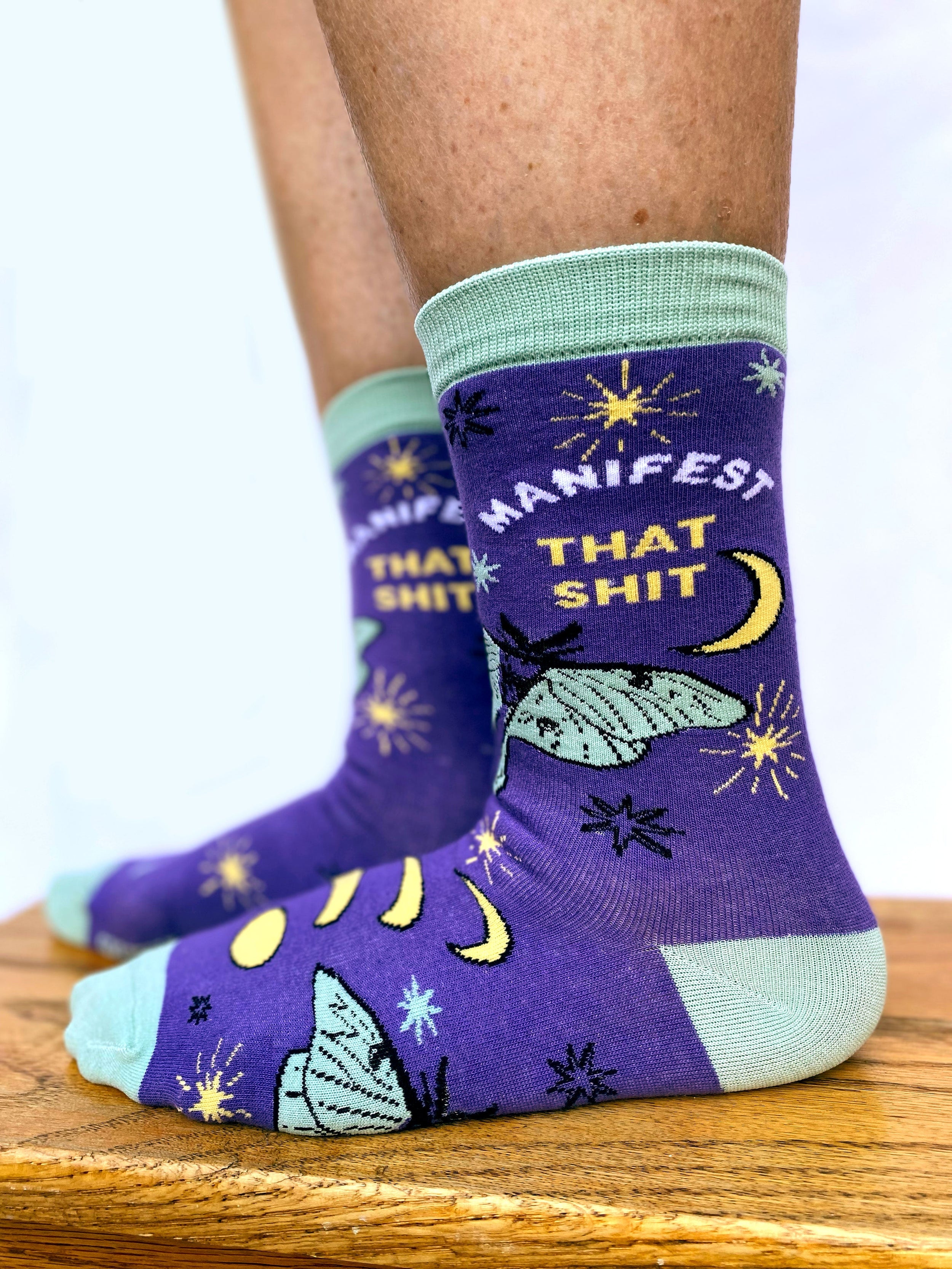 Model wearing a pair of socks with the slogan Manifest That S**t and a butterfly motif. Purple ankles, turquoise toe, heel and cuff.