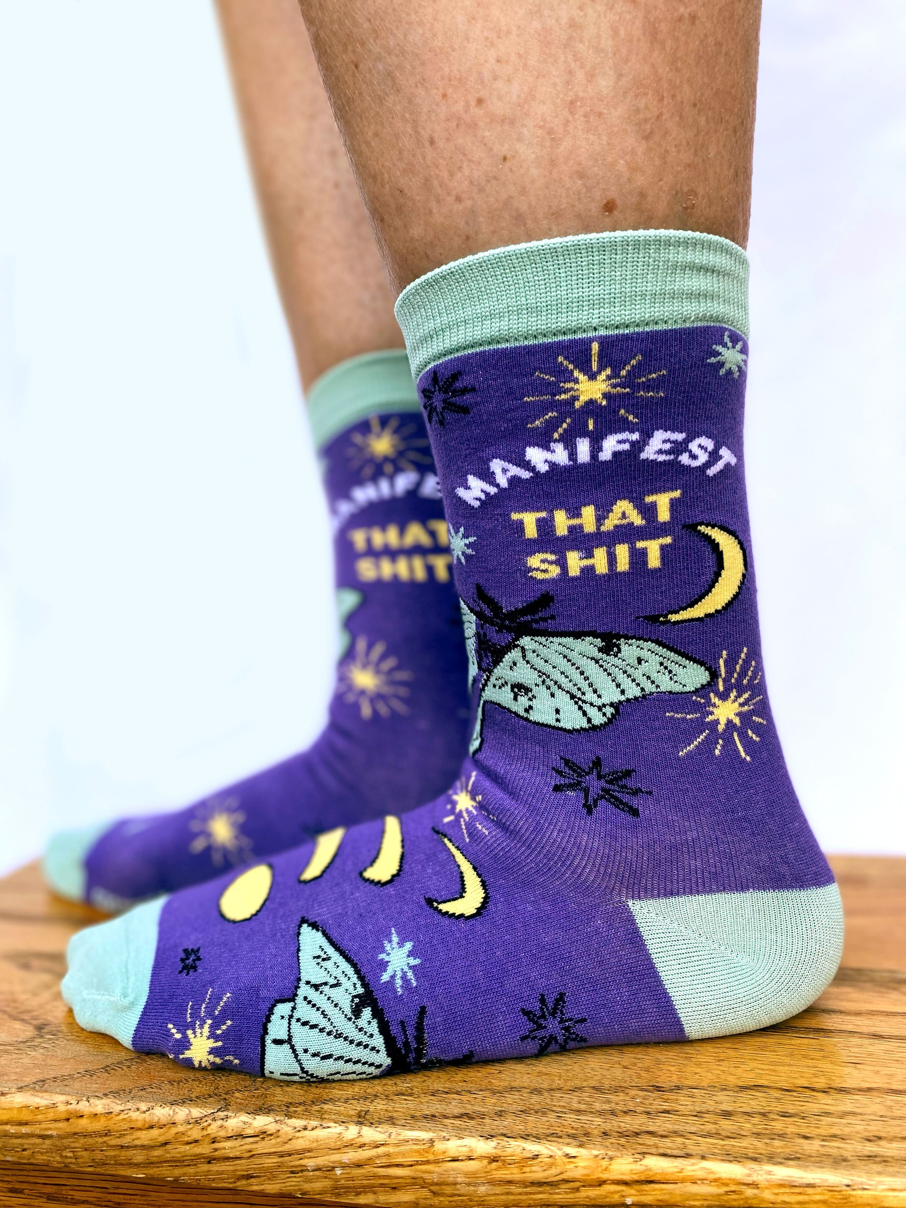 Model wearing a pair of socks with the slogan Manifest That S**t and a butterfly motif. Purple ankles, turquoise toe, heel and cuff.