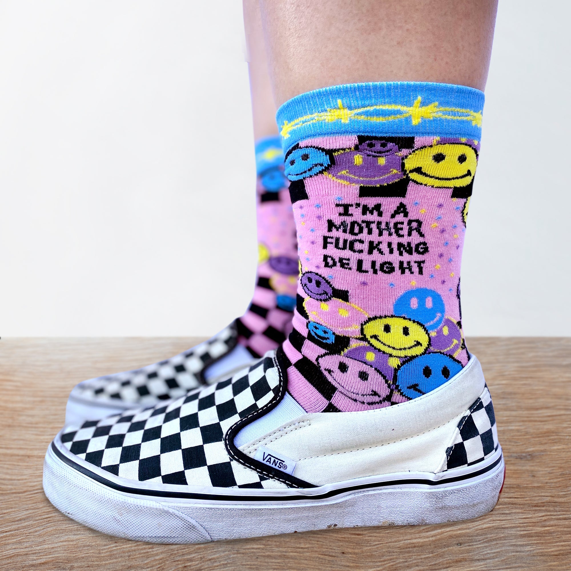 Model wearing a pair of socks with a bright pink and blue smiley face pattern and a bold sweary slogan. Blue toes, heel and cuff.
